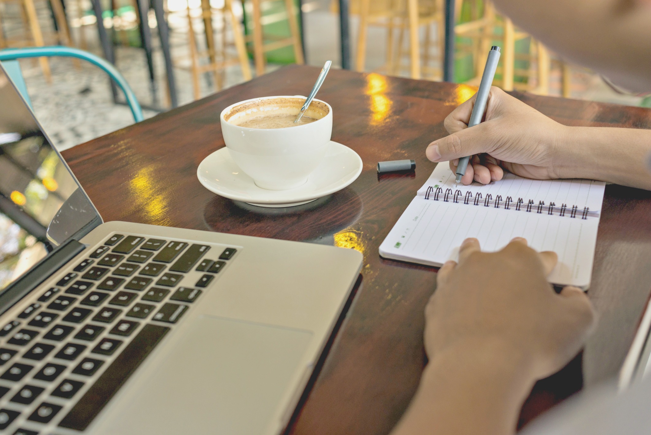 A person takes notes on a notepad with a laptop and coffee on a table